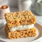 Rice Krispie Ice Cream – Nostalgic Crunch Meets Frozen Delight 5 A close-up of a rice krispie ice cream sandwich featuring a thick layer of creamy vanilla ice cream between two golden, crispy rice krispie treat layers. The sandwich is neatly stacked on a white plate with a blurred background of kitchen utensils and soft lighting.